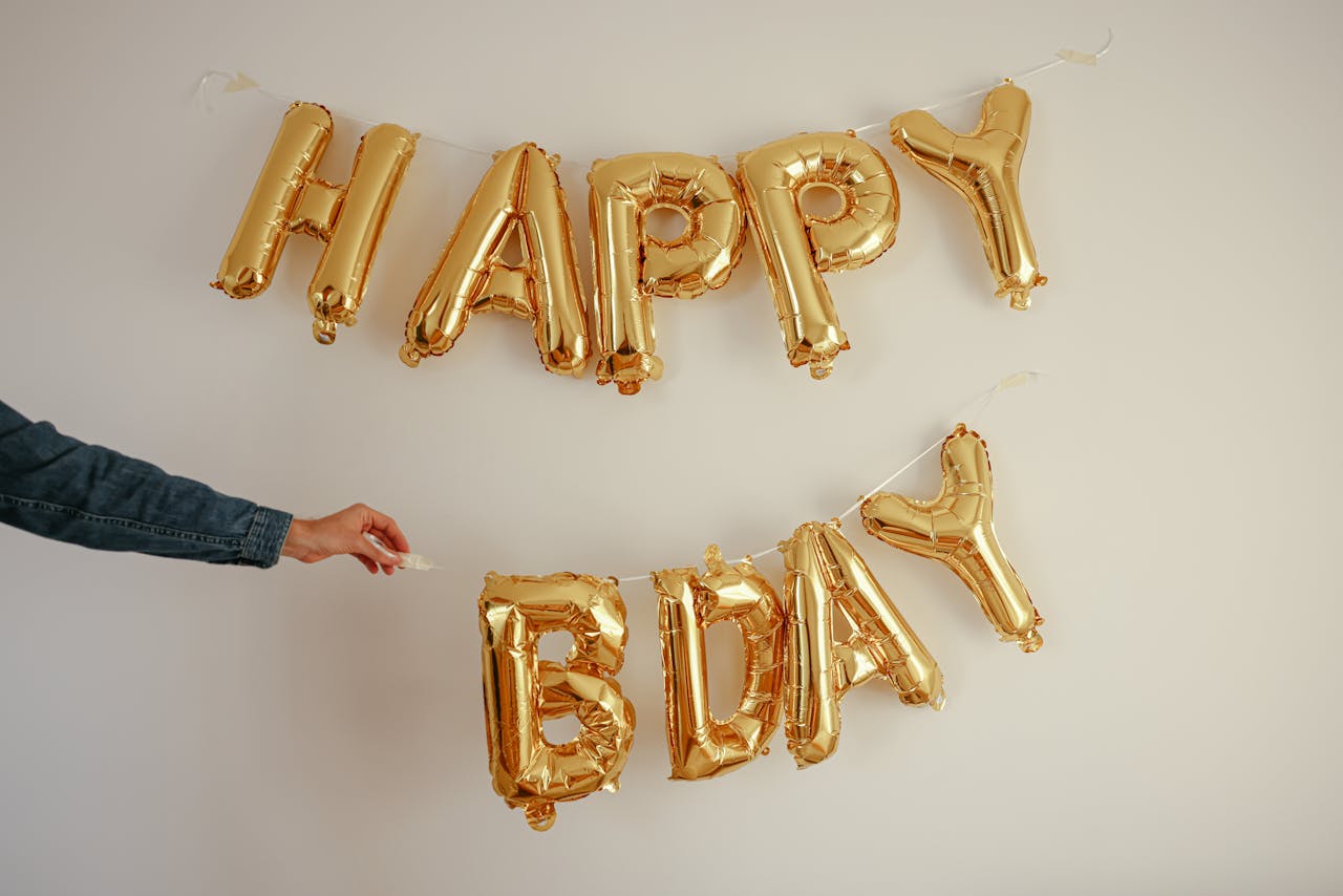 Golden foil balloons spelling 'Happy Bday' taped to a white wall with a hand adjusting them.